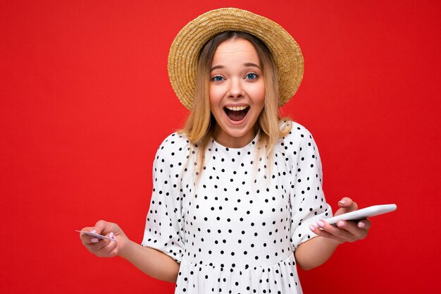 Foto di una donna bionda stupita e positiva attraente in abiti estivi che usa il cellulare e tiene in mano