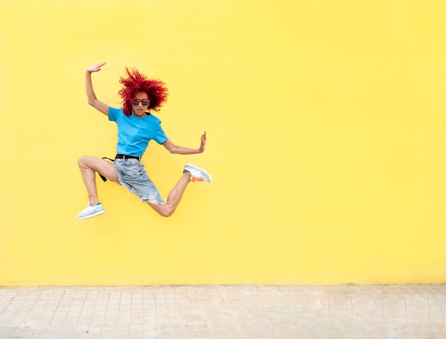 Foto di una donna con i capelli afro rossi che indossa una maglietta blu e occhiali da sole che salta su uno sfondo giallo