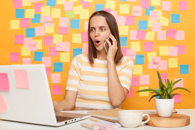 Foto di una donna dai capelli scuri scioccata che indossa una maglietta a righe seduta al tavolo in posa contro le schede memo sul muro giallo che lavora al computer portatile in ufficio parlando al cellulare tiene la bocca aperta