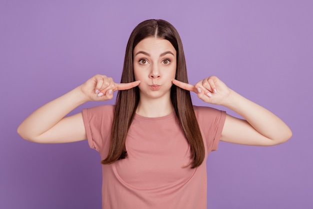 Foto di una donna divertente e infantile che punta le dita sulle guance gonfie isolate su sfondo viola