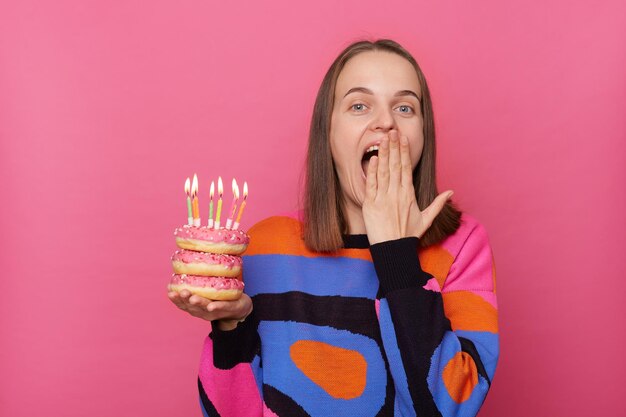 Foto di una donna eccitata e stupita con i capelli castani che indossa un elegante maglione che copre la bocca con il palmo che ha sorpresa per la sua festa di compleanno tenendo una ciambella con candele isolate su sfondo rosa