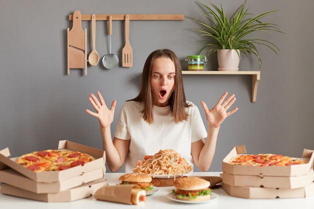 Foto di una donna eccitata stupita con i capelli castani che indossa una maglietta bianca seduta al tavolo con le braccia alzate e la bocca aperta pronta a mangiare pasta hamburger pizza e altro cibo spazzatura