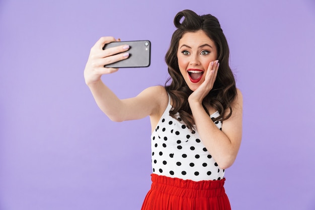 Foto di una donna pin-up glamour in abito vintage a pois che tiene e scatta una foto selfie su uno smartphone nero isolato su un muro viola