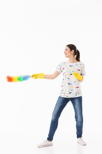 Foto di una donna positiva di 20 anni che indossa guanti di gomma gialli per la protezione delle mani che tiene in mano uno spolverino colorato mentre pulisce la stanza isolata su un muro bianco