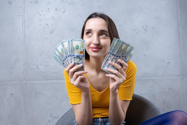 Foto di una donna ricca in abiti semplici con in mano un ventaglio di denaro dollaro isolato su uno sfondo di muro di cemento