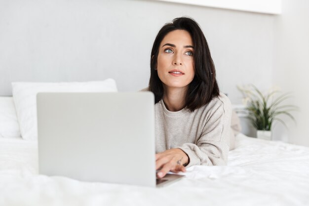 Foto di una donna soddisfatta 30s utilizzando laptop, mentre giaceva a letto con lenzuola bianche in una stanza luminosa