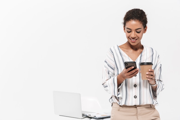 Foto di una giovane bella donna africana in posa isolata sul muro bianco usando il telefono cellulare che beve caffè.