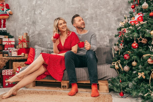 Foto di una giovane coppia accogliente e felice sera a casa, decorazioni natalizie, sfumature verdi rosse, donna e uomo con scatola regalo. Capodanno 2022. Tazza di tè o caffè. Gente felice