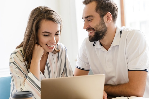 Foto di una giovane coppia felice di colleghi che lavorano con un computer portatile al chiuso in ufficio parlando tra loro.