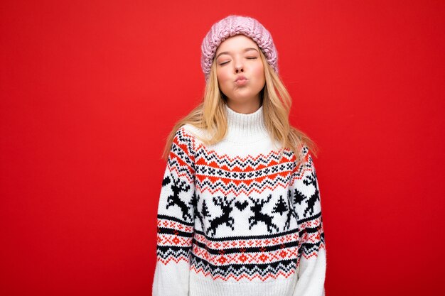 Foto di una giovane donna bionda abbastanza positiva e gioiosa isolata su una parete di fondo rossa che indossa un maglione invernale e un cappello rosa che dà un bacio