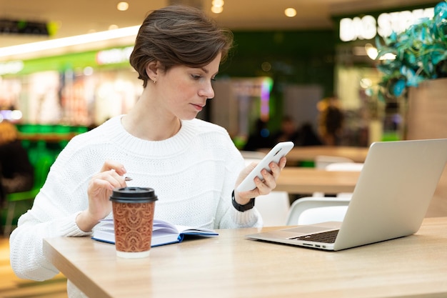 Foto di una giovane donna bruna attraente seduta in un centro commerciale a un tavolo con una tazza di carta da caffè e al lavoro su un computer portatile, utilizzando il telefono cellulare. Concetto di affari