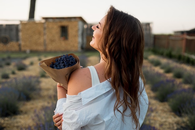 Foto di una giovane donna caucasica romantica in camicia bianca che tiene un mazzo di fiori mentre cammina all'aperto attraverso il campo di lavanda in estate