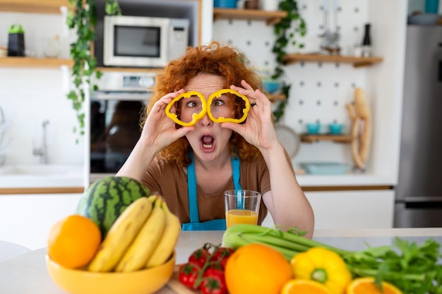 Foto di una giovane donna che sorride e tiene i cerchi di pepe sui suoi occhi mentre cucina insalata con verdure fresche nell'interno della cucina a casa