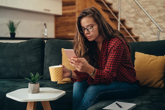 Foto di una giovane donna che usa lo smartphone mentre beve tè e si rilassa a casa