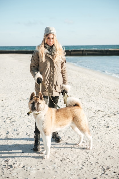 Foto di una giovane donna concentrata che cammina sulla spiaggia invernale con un cane al guinzaglio.