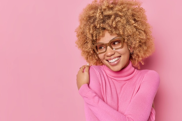 Foto di una giovane donna di bell'aspetto che si abbraccia tocca le spalle sorride teneramente indossa gli occhiali e il dolcevita casual sembra felicemente lontano isolato su sfondo rosa. Autostima