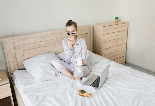 Foto di una giovane donna gioiosa in pigiama che digita sul laptop e sorride mentre è seduta sul letto in una stanza luminosa