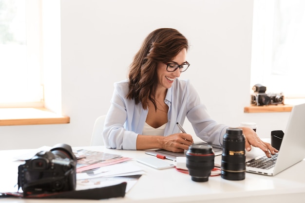 Foto di una giovane fotografa sorridente allegra in ufficio che lavora con un computer portatile e una tavoletta grafica.