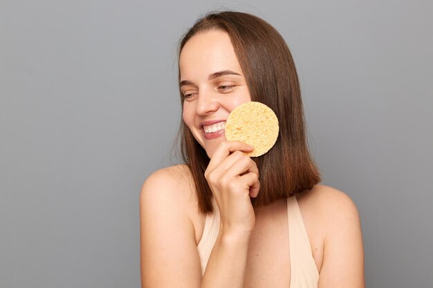 Foto di una giovane ragazza adulta felice con un sorriso gentile che tiene una spugna cosmetica vicino al viso isolato su sfondo grigio che distoglie lo sguardo godendo di procedure cosmetiche