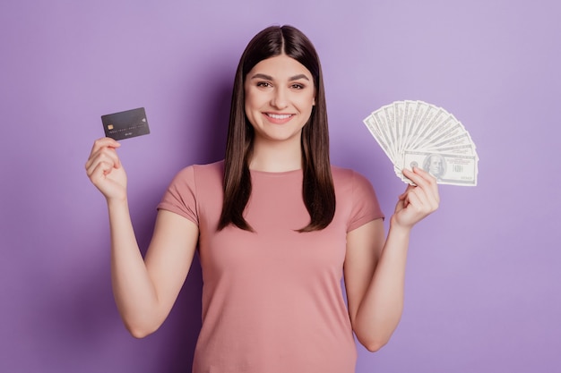Foto di una giovane ragazza attraente e felice, sorriso positivo che tiene in mano una carta di credito dollari in contanti, un ricco sfondo di colore viola isolato