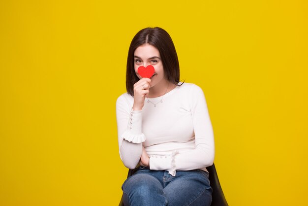 Foto di una giovane ragazza sorridente allegra si nasconde il viso dietro un cuore rosso su spazio giallo.