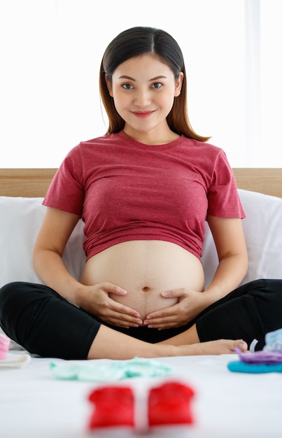 Foto di una graziosa scarpetta rossa su un letto bianco con un focus morbido su una donna incinta seduta su un letto con cose per bambini. Aspettando un bambino sano e un concetto di giorno del parto.