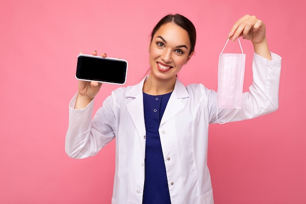 Foto di una persona di sesso femminile adulta sorridente che indossa un camice bianco che tiene in mano una maschera che mostra un cellulare moderno