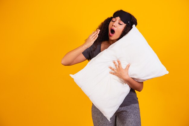 Foto di una ragazza afro che abbraccia un cuscino su sfondo giallo che sbadiglia dal sonno.