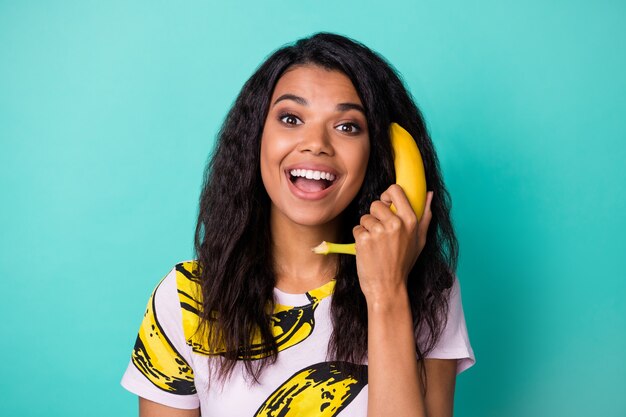 Foto di una ragazza afroamericana positiva che chiama telefono banana indossa una maglietta rosa isolata su uno sfondo color verde acqua