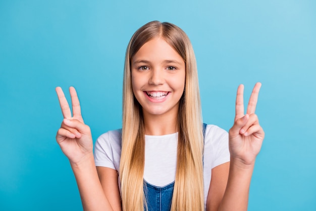 Foto di una ragazza bionda positiva che mostra il v-sign nella telecamera indossa una maglietta bianca isolata su uno sfondo di colore blu pastello