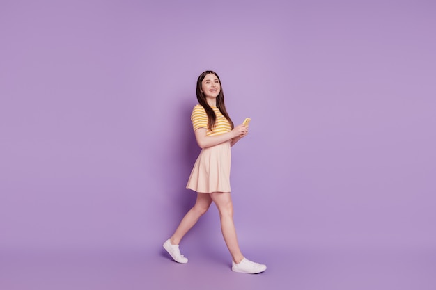 Foto di una ragazza carina affascinante e positiva che cammina tenendo il telefono in chat su sfondo viola
