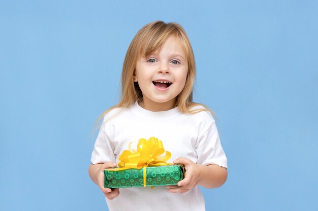 Foto di una ragazza carina e dolce che indossa una maglietta bianca che sorride con in mano una scatola regalo gialla e verde isolata sullo sfondo di colore blu