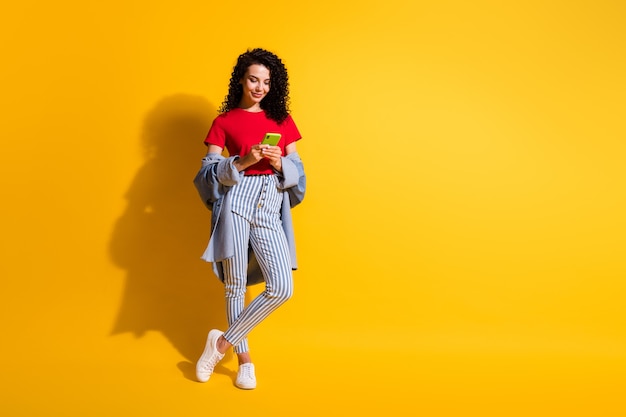 Foto di una ragazza che tiene il telefono guarda lo schermo indossa jeans a righe t-shirt rossa giacca scarpe da ginnastica isolate sfondo di colore giallo