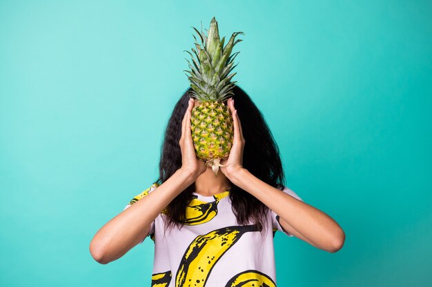 Foto di una ragazza dalla pelle scura che copre il viso con un ananas ecologico isolato su uno sfondo di colore turchese