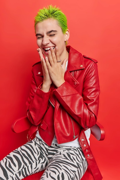 Foto di una ragazza hipster felicissima con i capelli verdi alla moda che ride di qualcosa vestito con giacca di pelle e leggings a righe isolati su sfondo rosso vivo. Concetto di persone ed emozioni positive