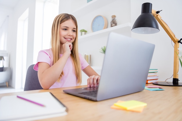 Foto di una ragazza positiva che si siede al tavolo, studia, usa il computer a distanza, leggi, guarda, video, webinar, tocca la mano, il mento in casa, al chiuso