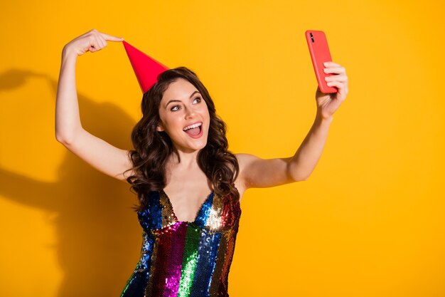 Foto di una ragazza stupita blogger influencer prendere selfie smartphone puntare il dito il suo cono rosso copricapo indossare gonna lucida arcobaleno isolata su sfondo di colore brillante brillante