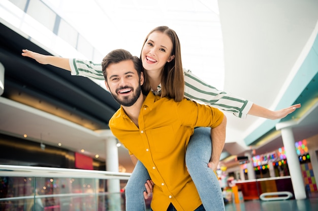 Foto di una signora carina allegra che allarga le braccia come ali bel ragazzo che la porta sulle spalle visita il centro commerciale del centro commerciale insieme buon umore divertendosi a giocare indossando abiti casual al chiuso