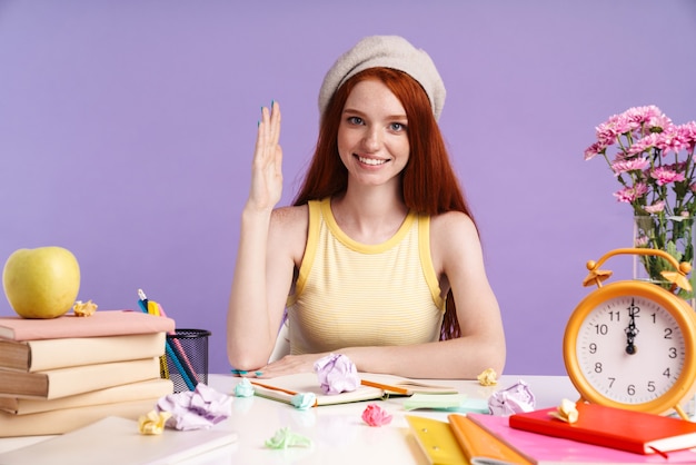 Foto di una studentessa rossa che alza la mano mentre è seduta alla scrivania a lezione con quaderni isolati su un muro viola