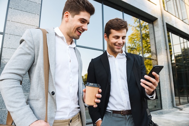 Foto di uomini d'affari europei in giacca e cravatta che utilizzano smartphone, mentre in piedi all'aperto vicino a un edificio con caffè da asporto