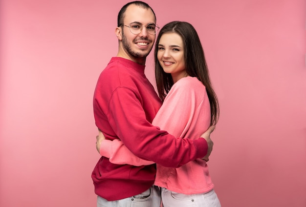 Foto di uomo attraente con barba in abiti rossi e donna in rosa si abbracciano e sorridono. La coppia sembra felice, isolata su sfondo rosa.