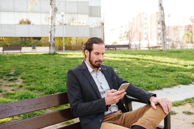 Foto di uomo attraente intelligente 30s in tuta da lavoro che tiene e utilizza lo smartphone, mentre è seduto su una panchina nel parco cittadino durante la giornata di sole