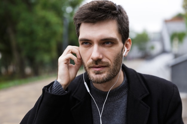 Foto di uomo caucasico 20s che indossa gli auricolari utilizzando il telefono cellulare, mentre si cammina all'aperto