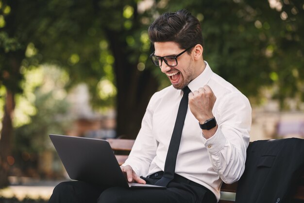 Foto di uomo d'affari bello in vestito che si siede sulla panchina nel parco verde e lavora al computer portatile durante la giornata di sole