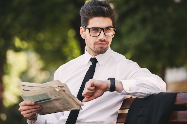 Foto di uomo d'affari serio in tuta leggendo il giornale su una panchina nel parco verde e guardando l'orologio da polso