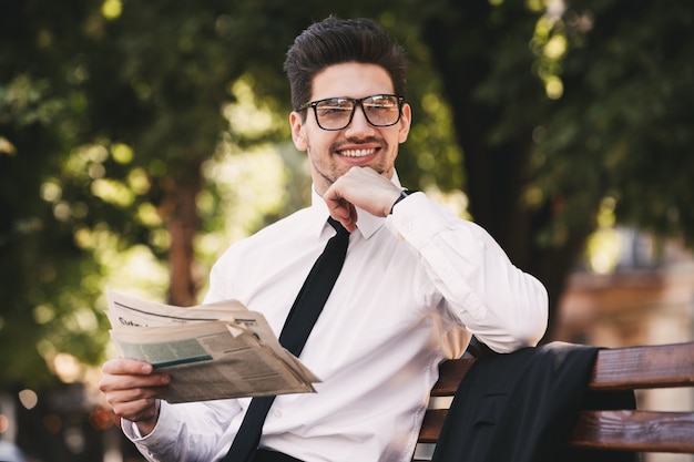 Foto di uomo felice in giacca e cravatta seduto su una panchina nel parco verde e leggere il giornale durante la giornata di sole