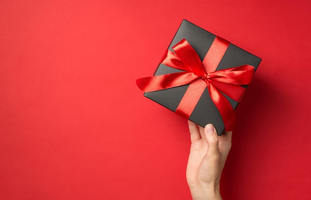 Foto di vista dall'alto in prima persona della mano della ragazza che tiene una confezione regalo nera con fiocco di nastro rosso su sfondo rosso isolato con spazio vuoto