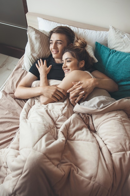 Foto di vista superiore di una coppia caucasica sdraiata sul letto che si abbraccia sotto un copriletto e sorridere in una mattina