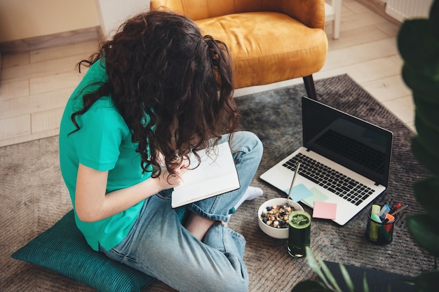 Foto di vista superiore di uno studente che fa i compiti sul pavimento e utilizza un laptop mentre mangia cereali con succo di verdura