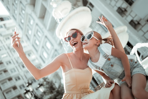 Foto divertenti. Madre e figlia gioiose sedute per strada e che giocano alla scimmia facendo selfie sullo smartphone.